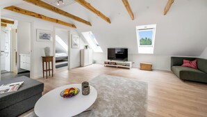 Een ruime loft met zichtbare houten balken, een flatscreen televisie, een groene bank en een eethoek met een witte tafel.