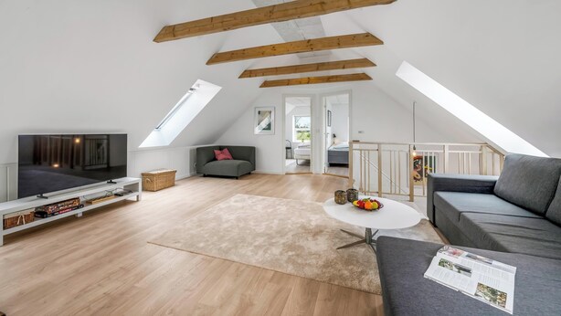 Een ruime loft met hoge plafonds, zichtbare houten balken, een grote televisie, een grijze bank, een klein rond tafeltje en een trap naar een hoger gelegen verdieping.