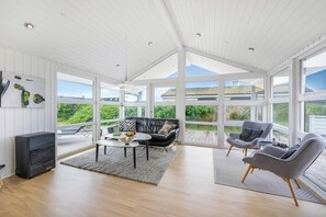 A spacious room with a large window, a black leather sofa, two grey armchairs, a small table, and a fireplace. - Lovely holiday home with a wilderness bath, close to the North Sea and. (Ringkøbing)