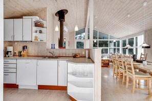 A modern kitchen with white cabinetry, a central island, and a dining area with wooden chairs. - Bright holiday home with electric charger, terrace and wood-burning stove (Fjerritslev)