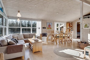 A spacious living area with a sofa, dining table, chairs, and a large window. - Bright holiday home with electric charger, terrace and wood-burning stove (Fjerritslev)