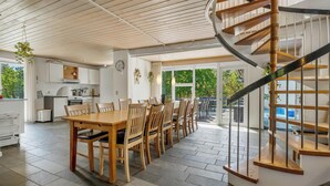 A modern kitchen with white cabinets, a dining area with wooden tables and chairs, and a spiral staircase. - Lovely 16-person pool house with spa, sauna and large terrace (Vestervig)