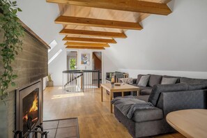 A loft-style room with a fireplace, wooden beams, a gray sofa, and a dining table. - Lovely 16-person pool house with spa, sauna and large terrace (Vestervig)