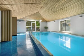An indoor swimming pool with a wooden ceiling, clear glass doors leading outside, and a potted plant on the right. - Lovely 16-person pool house with spa, sauna and large terrace (Vestervig)