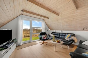 En moderne stue med fjernsyn, en sort lædersofa, en sort lænestol, et lille hvidt bord og et stort vindue med udsigt til det fri.
