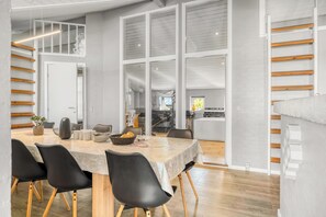 A modern dining area with a large table, black chairs, and a view of the kitchen and upper floor. - Renovated holiday home with hot tub, sauna and shelter (Vestervig)