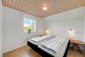 Ein Zimmer mit schrägem Dach, einem Fenster mit Blick auf Grünflächen, zwei Betten mit weißer Bettwäsche, einem Nachttisch und einer wandmontierten Leuchte.