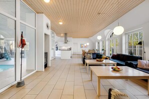 A spacious living area with a kitchen in the background, large windows, and a pool visible through the glass doors. - Large holiday home with pool, hot tub and activity room in Husby (Ulfborg)