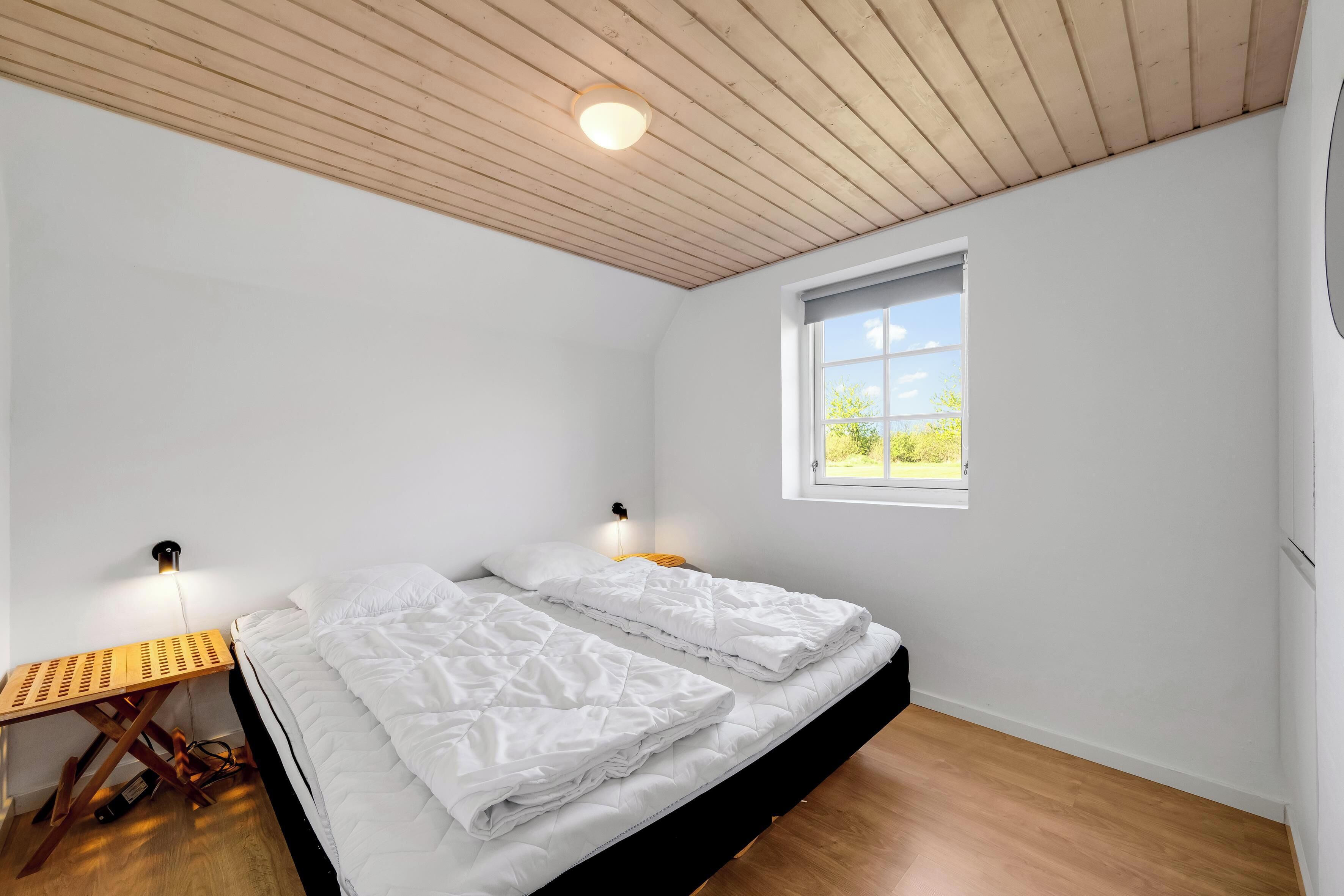 Een slaapkamer met een bed, nachtkastje, lamp, raam en een houten plafond.