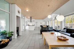A modern kitchen with white cabinets, a central island, and a dining area with a black leather sofa and wooden table. - 18-person pool house with hot tub, infrared sauna and activity room. (Ulfborg)