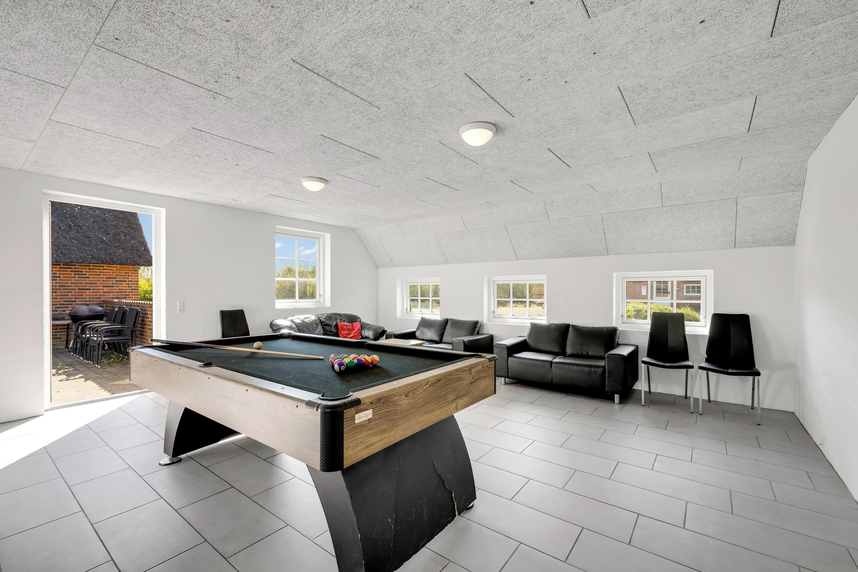 Una mesa de billar moderna en el interior, con tapa de madera y patas negras, rodeada de sillas negras y un sofá en una habitación luminosa con ventanas amplias.