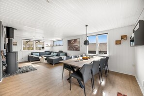 A modern living room with a sofa, dining table, and chairs, a fireplace, and large windows. - Holiday home with enclosure for dogs, 500 meters from the North Sea (Ringkøbing)
