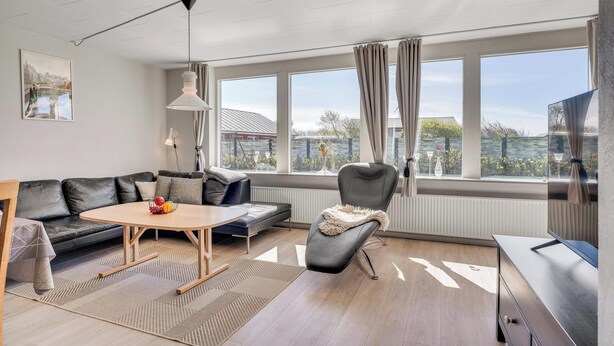 A modern living room with a sofa, a round table with fruit, a chair with a fur throw, a TV, and large windows with curtains. - 14-person holiday home in Agger with wilderness bath and outdoor sauna (Vestervig)