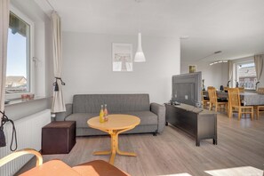 Ein modernes Wohnzimmer mit einem grauen Sofa, einem runden Couchtisch, einer TV-Konsole und einem Essbereich mit Holzsesseln und einem Tisch.