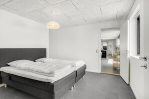 A modern bedroom with a large bed, a hanging light fixture, and a view into the dining area. - Renovated holiday home with enclosed terrace, centrally located in Vorupør (Thisted)