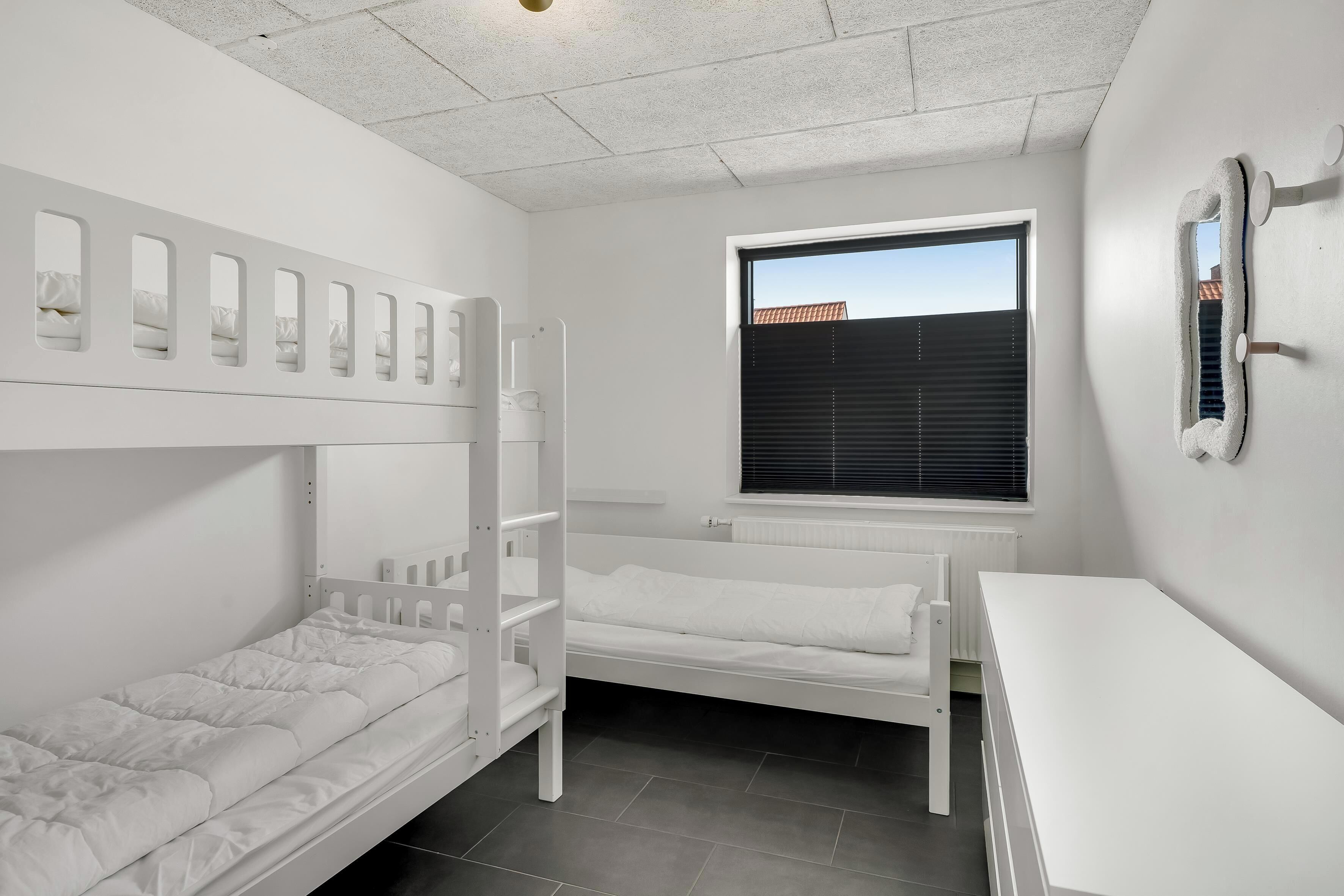 A room with bunk beds, a window with blinds, a white desk, and a mirror on the wall.