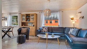 Ein modernes Wohnzimmer mit einem Ecksofa, einem runden Couchtisch, einem Essbereich mit Holztisch und -stühlen sowie einer Küchenzeile mit Schränken und einem Fenster.