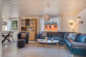 Un salon moderne comprenant un canapé d’angle, une table basse ronde, un coin repas avec une table et des chaises en bois, et un coin cuisine avec des placards et une fenêtre.