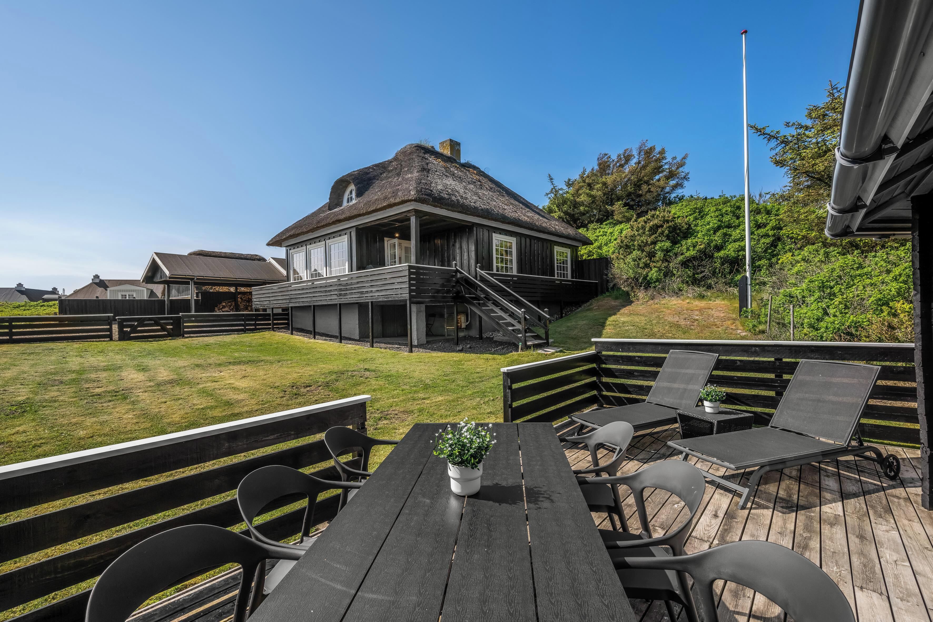 Een huis met rieten dak, een trap naar een terras, buitenzitplaatsen met stoelen en tafels, en een vlaggenmast.