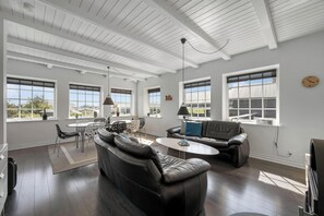 A spacious living area with a high ceiling, large windows, a sofa, a coffee table, and a dining set. - Charming modernized holiday home in the heart of Søndervig (Ringkøbing)