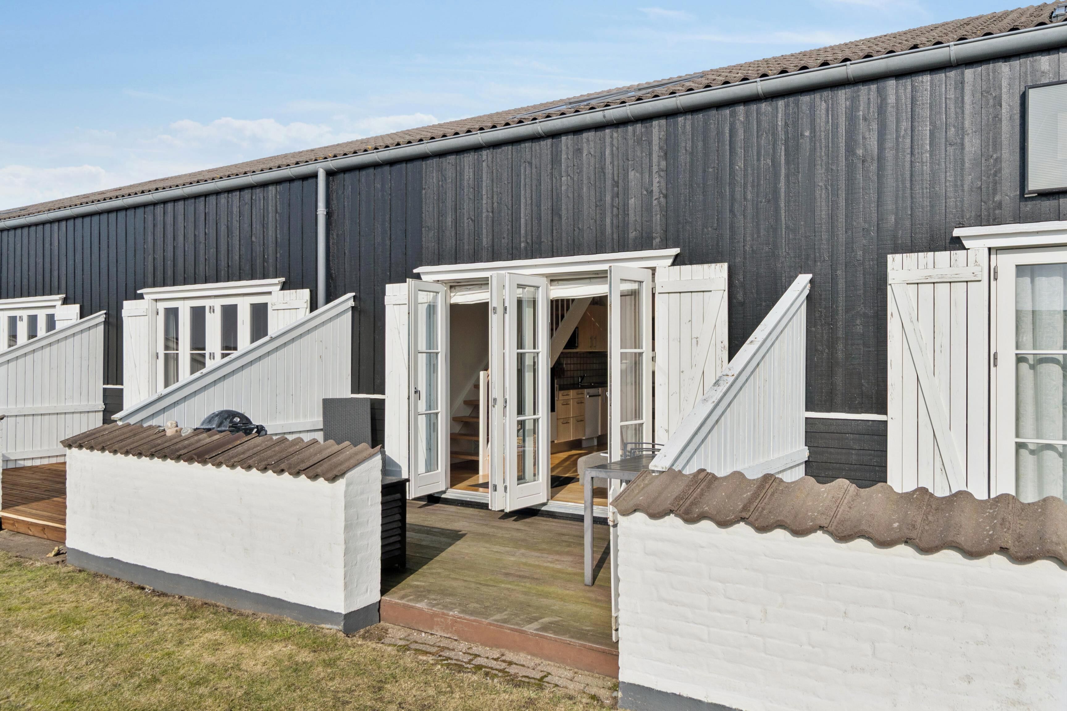 Een rij witte blokhutten met openslaande deuren, een houten terras, en een grijs gebouw op de achtergrond.