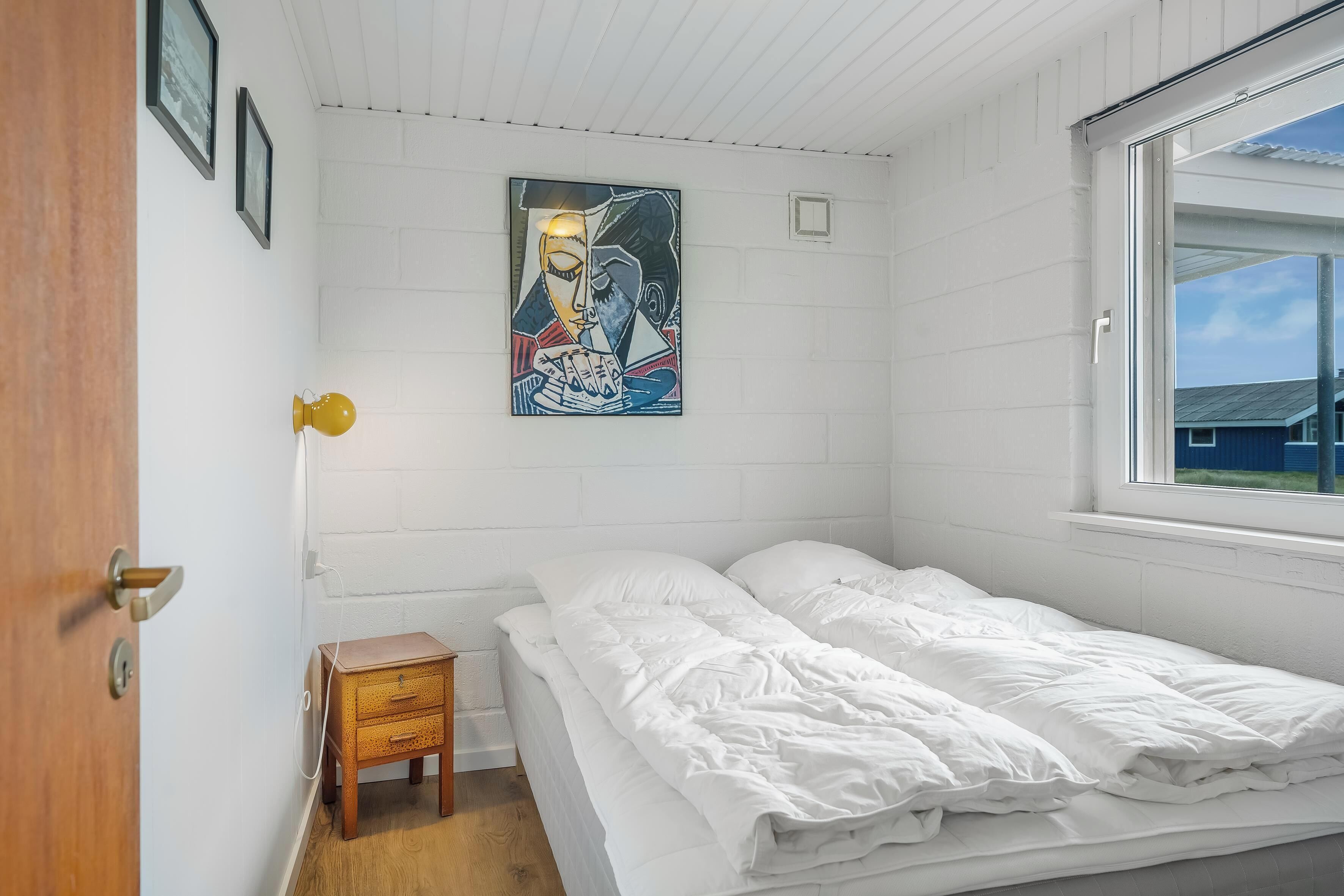 Een slaapkamer met een bed, een klein houten nachtkastje, een aan de muur hangend schilderij en een raam.