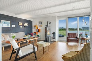 A modern living room with a fireplace, wooden furniture, and a large window offering a view of the outdoors. - Lovely holiday home with large terrace close to the North Sea in Klitmøller (Thisted)