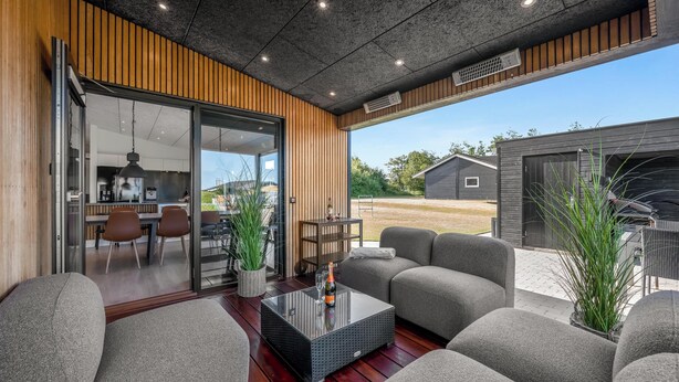 A modern outdoor living area with a glass door leading to a kitchen, a black coffee table with a bottle, and grey sofas. - Lovely holiday home with mini golf course, sauna, wilderness bath and view of. (Vinderup)
