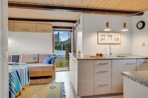 Eine kleine Küche mit Spüle, Arbeitsfläche und Schränken, ein Essbereich mit Tisch und Sofa, und ein Blick durch eine offene Tür ins Freie.