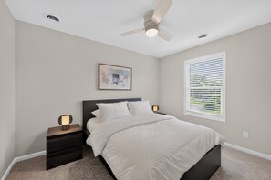 Une chambre à coucher comprenant un lit, des tables de chevet, un ventilateur de plafond, une fenêtre avec des stores et une œuvre d’art accrochée au mur.