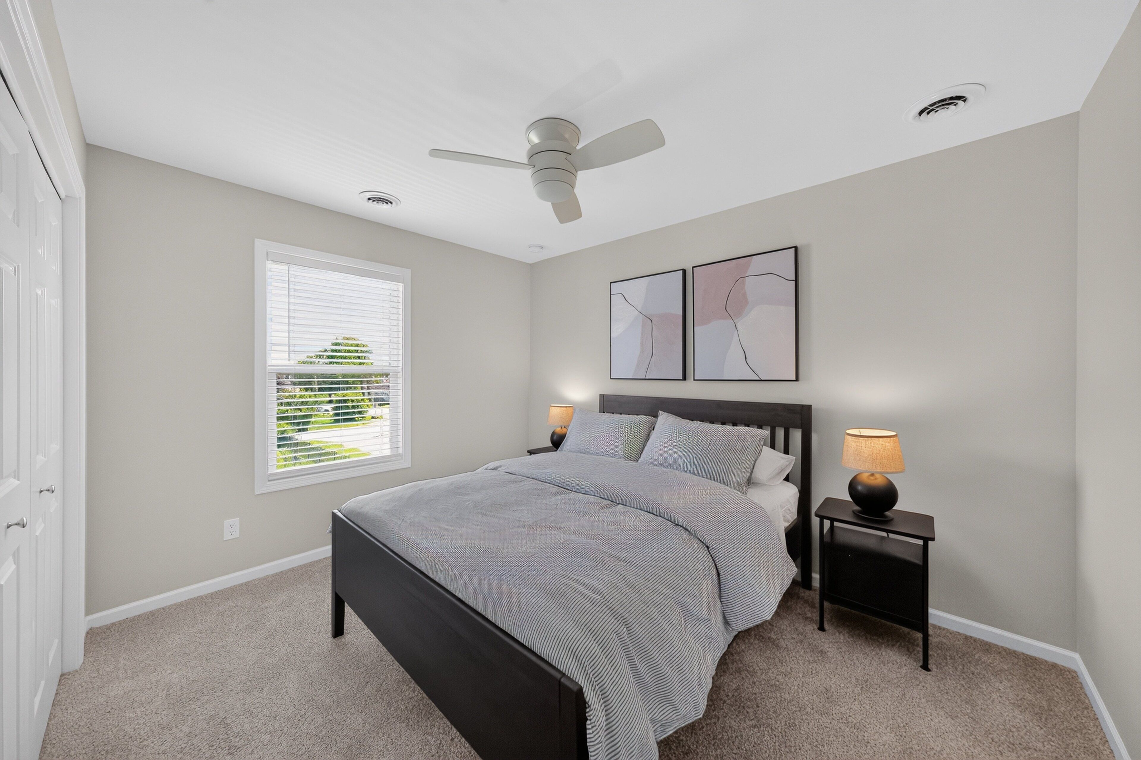 Une chambre à coucher avec un lit, un ventilateur de plafond, une table de chevet avec une lampe, une fenêtre avec des stores et des œuvres d’art accrochées au mur.