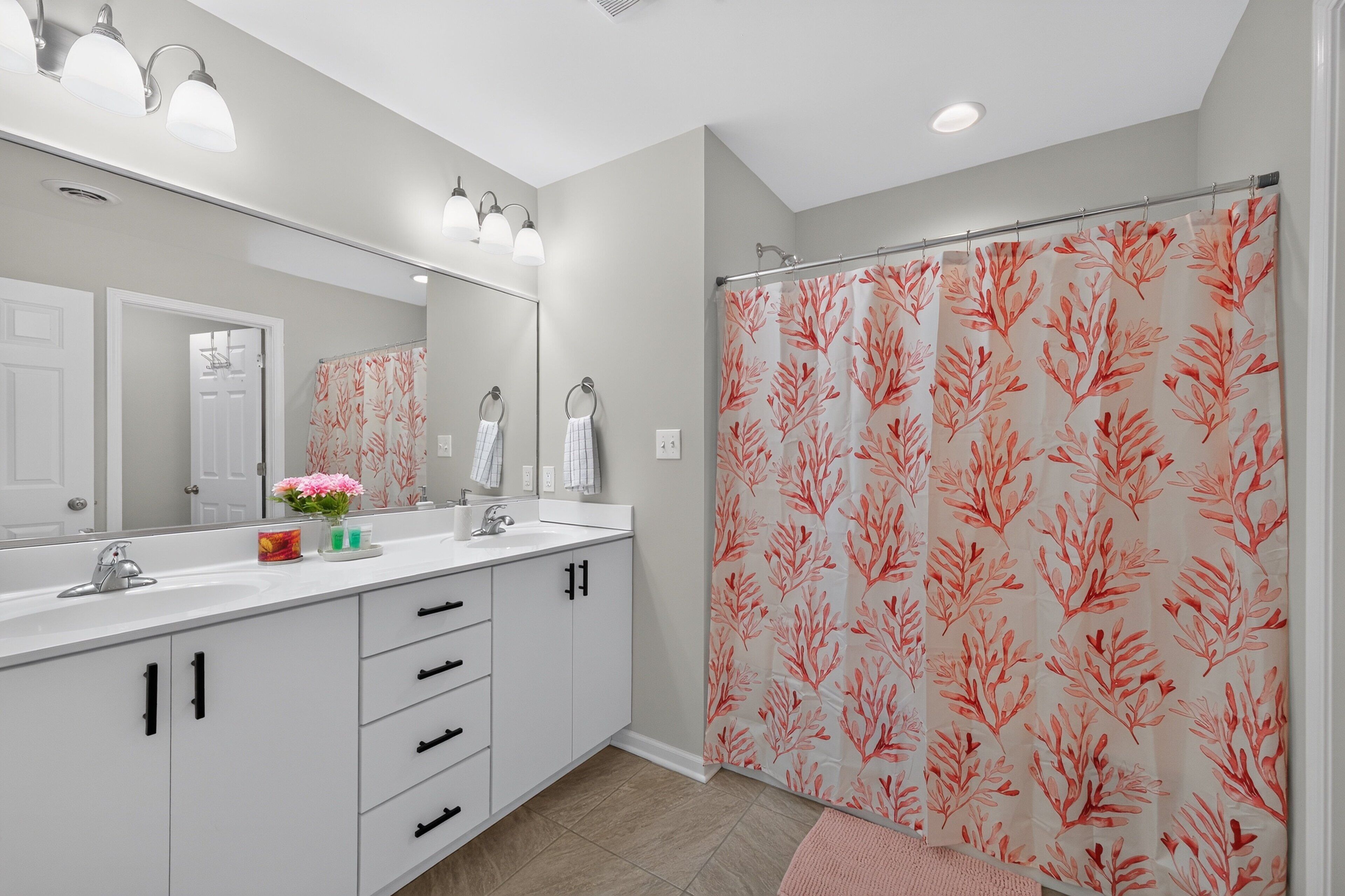 A bathroom with a white vanity, a large mirror, a shower curtain with a leaf pattern, and a pink rug.