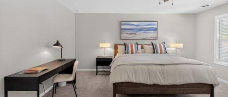 A bedroom with a bed, desk, chair, two bedside tables with lamps, a ceiling fan, and a painting on the wall.
