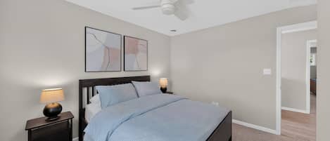 Un dormitorio con una cama, dos mesitas de noche con lámparas, un ventilador de techo y dos obras de arte colgadas en la pared.