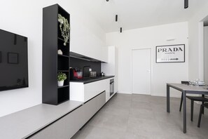 Eine moderne Küche mit weißen Schränken, einer schwarzen Regaleinheit mit Pflanzen, einer Kaffeemaschine und einem Essbereich mit Tisch und Stühlen.