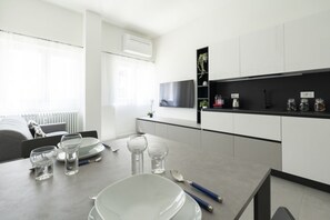 A modern kitchen with white cabinets, a black countertop, and a dining area with a table set for two. - GuestHost - Comfy TERRA Apartment near Cenisio MM5 (Milan)