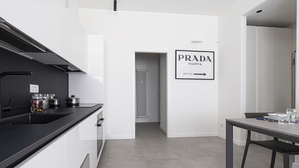 A modern kitchen with a black countertop, white cabinets, and a dining area with a table and chairs. - GuestHost - Comfy TERRA Apartment near Cenisio MM5 (Milan)