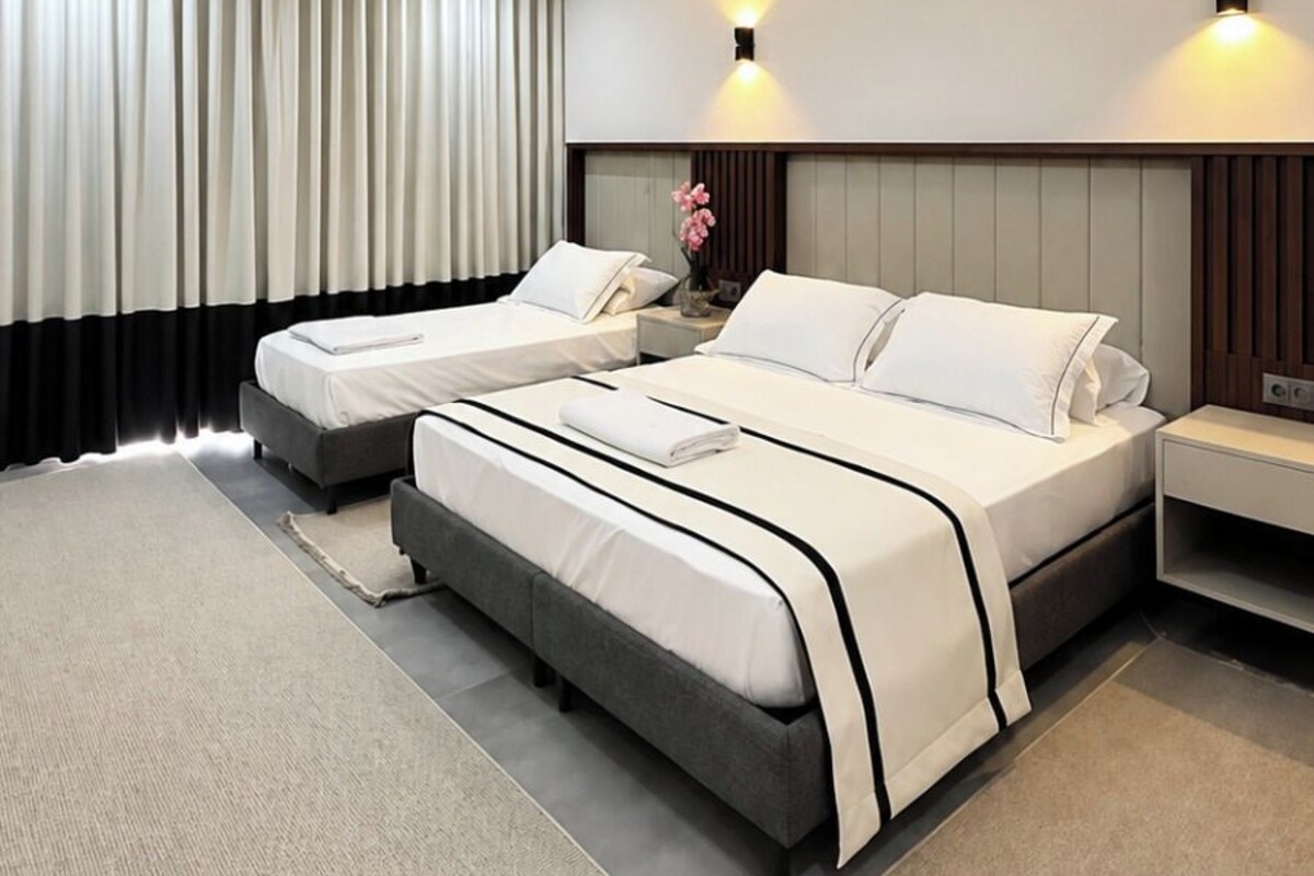 Una habitación de hotel moderna con dos camas, un ventanal con cortinas, un televisor de pantalla plana y una mesita de noche con lámpara.