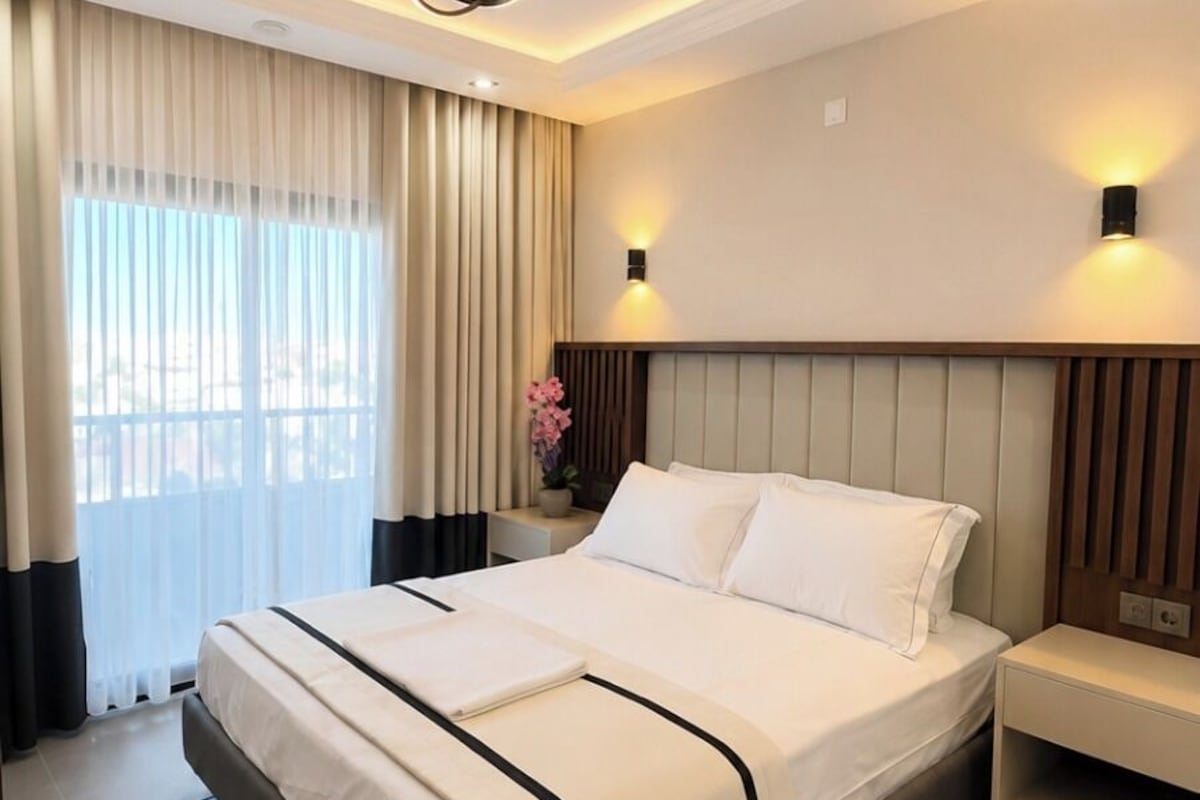 Una habitación de hotel moderna con una cama grande, mesitas de noche, una lámpara de pared y una ventana con cortinas.