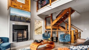 Ein modernes Wohnzimmer mit Kamin, blauen Sesseln, einer Holztreppe und einem großen Fenster mit Blick auf die Berge.