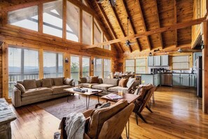A spacious living area with a wooden ceiling and large windows, a kitchen with light-colored cabinets, and a cozy seating arrangement. - Two cabins - Sleeps 24 - Views - Games - Outdoors (Sevierville)