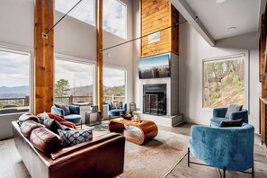 A modern living room with a fireplace, large windows, and a view of the outdoors. - Two cabins - Sleeps 24 - Views - Games - Outdoors (Sevierville)