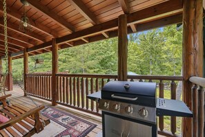 Eine überdachte Terrasse im Freien mit Grill, Schaukelstühlen aus Holz und Blick auf einen Wald.