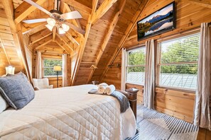 Ein Schlafzimmer einer Holzhütte mit Bett, Deckenventilator, Fenstern und einem Fernseher.