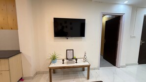 Ein Wohnzimmer mit einem an der Wand montierten Flachbildfernseher, einem kleinen Tisch mit einer Pflanze, einem gerahmten Bild und einer Tür zu einem anderen Raum.