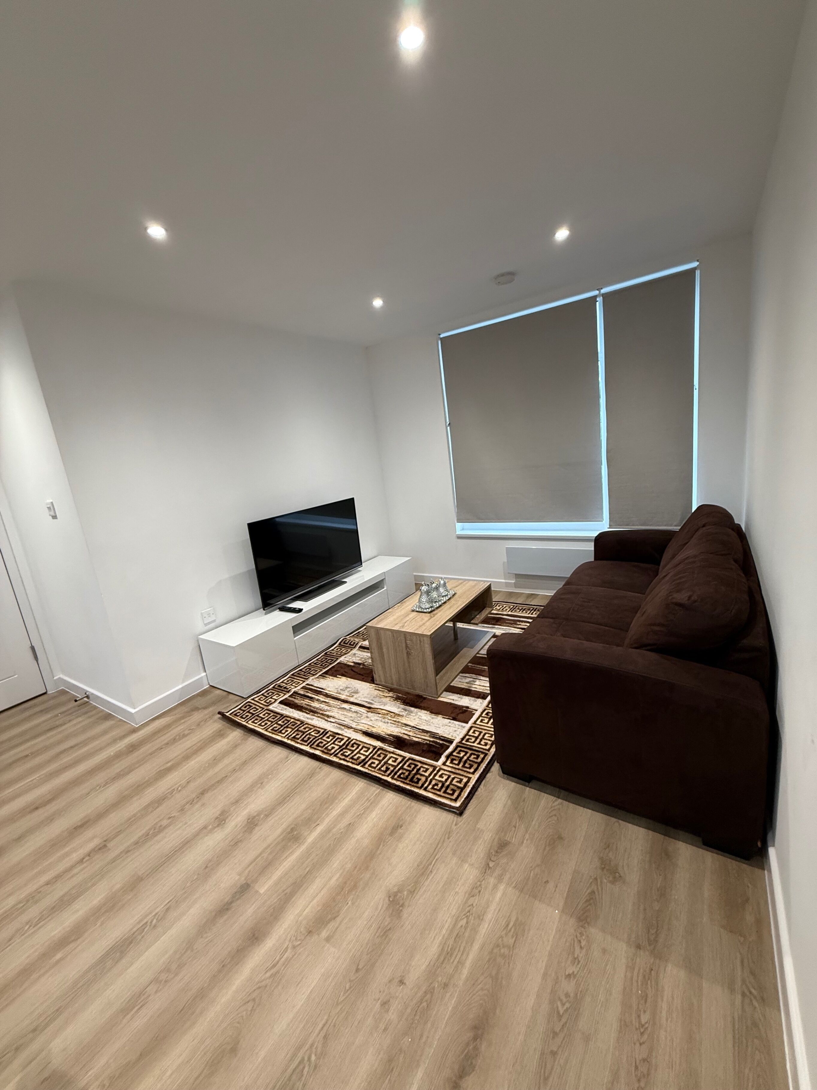 Ein modernes Wohnzimmer mit Fernseher, braunem Sofa, Couchtisch aus Holz und einem gemusterten Teppich.