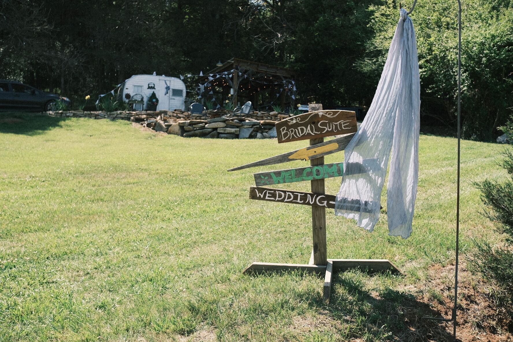 Un panneau indicateur en bois, sur lequel on peut lire les mots « LIEU DU MARIAGE » et « CÉRÉMONIE », est installé dans une zone herbeuse, avec une tente blanche en arrière-plan.