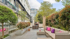 Een buitenplaats met zitgedeeltes, plantenbakken en een uitzicht op een modern gebouw op de achtergrond.