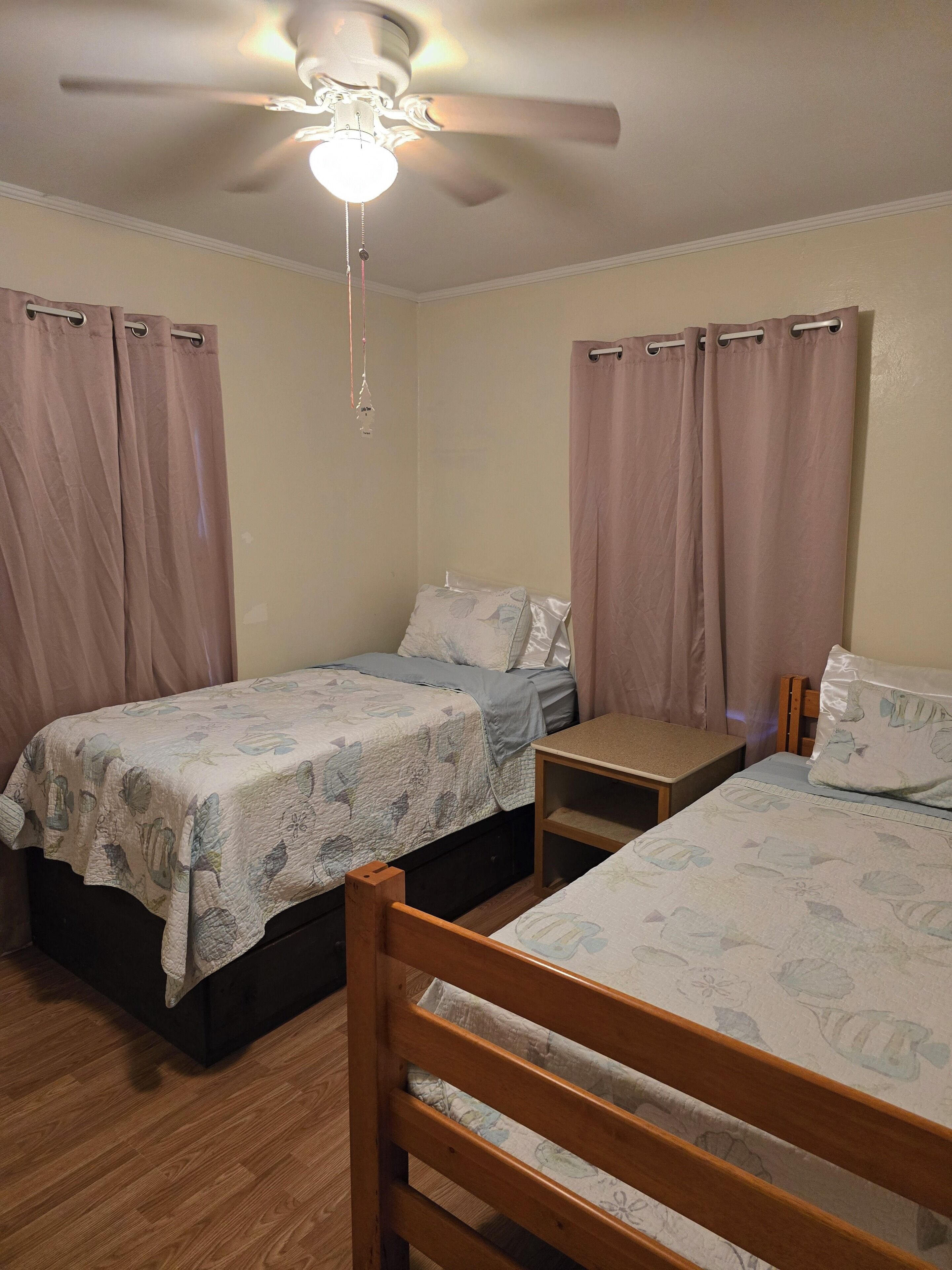 A bedroom with two single beds, a wooden headboard, a nightstand, a ceiling fan, and curtains.