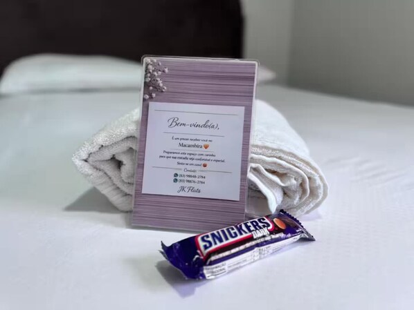 A neatly folded white towel, a card with a message, and a Snickers chocolate bar on a bed.